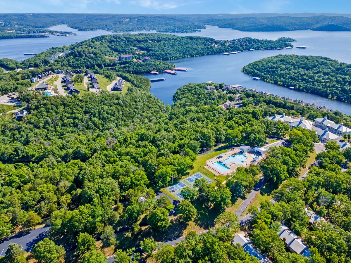 This wide-angle view captures the resort swimming pool, tennis courts, lush greenery, and the sparkling lake with surrounding hills in the backdrop. The layout showcases a variety of resort amenities in one cohesive scene, including recreation spaces and natural beauty.