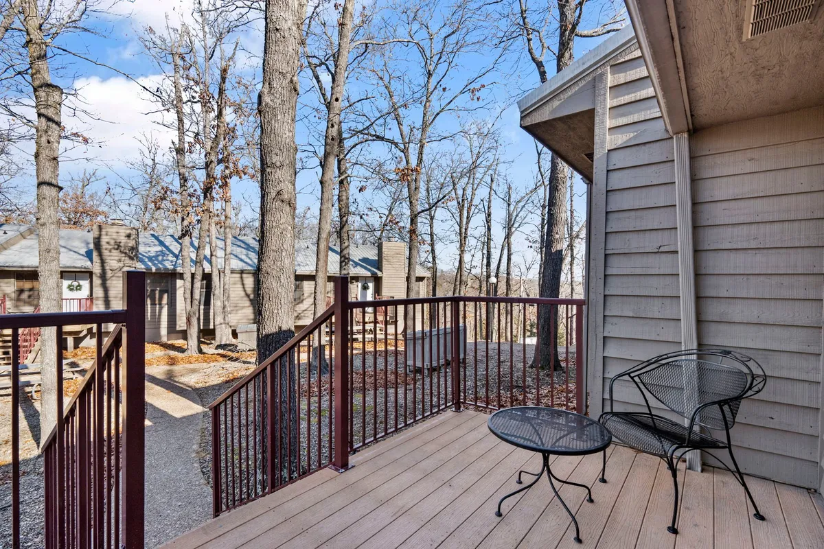 A cozy wooden deck featuring a small table and chairs, surrounded by lush green trees. The intimate arrangement suggests a perfect space for enjoying coffee, reading, or quiet conversations, with natural light filtering through the foliage.