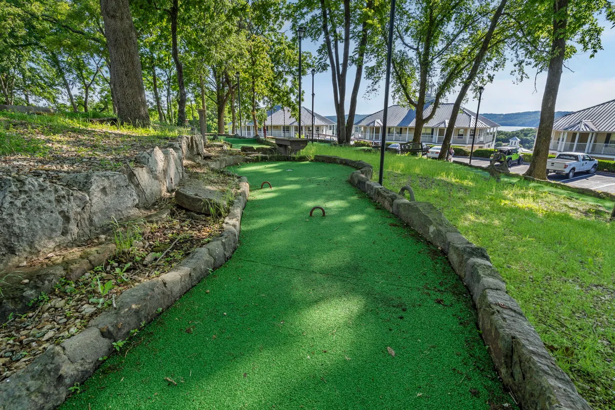 A vibrant mini-golf pathway winds through green turf with natural stone borders and is surrounded by tall trees, creating a shaded and playful atmosphere. The area is clearly designed for outdoor leisure and family fun, encouraging friendly competition and group activities in a lush setting.