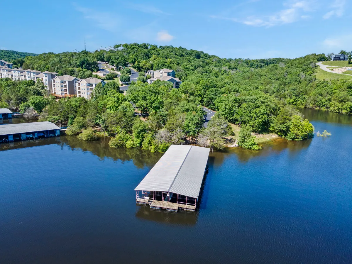 The tranquil lake scene features a dock extending out over the water, encircled by lush greenery and flanked by residential buildings. It emphasizes easy access to lakeside recreation like boating and fishing.