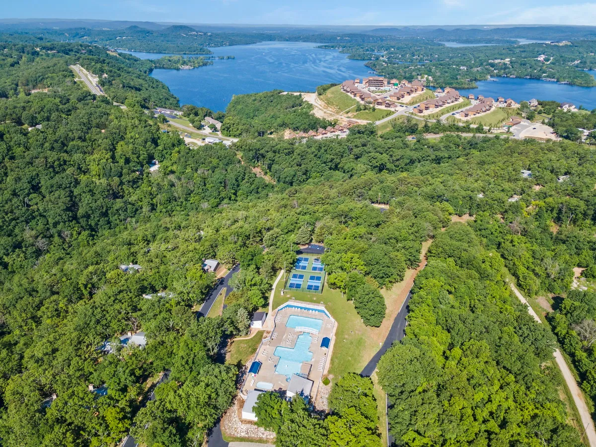 A panoramic view showcases a large swimming pool nestled among green trees, with Table Rock Lake visible nearby and residential areas dotting the hillsides. The scene evokes a sense of tranquility and connection to both the water and the surrounding natural beauty.