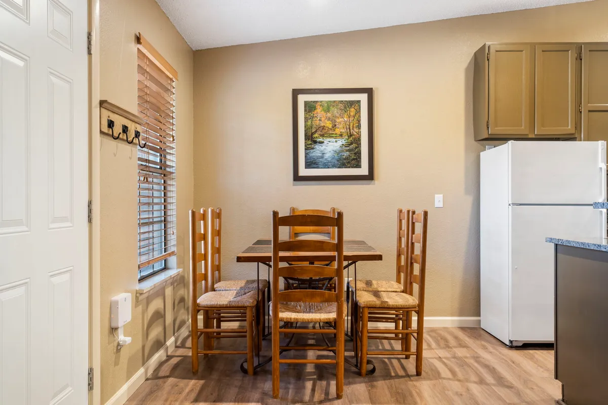 A cozy dining area featuring a wooden dining table, framed artwork, and a refrigerator in the background. The setup offers a perfect spot for family meals and casual gatherings, surrounded by comforting décor.