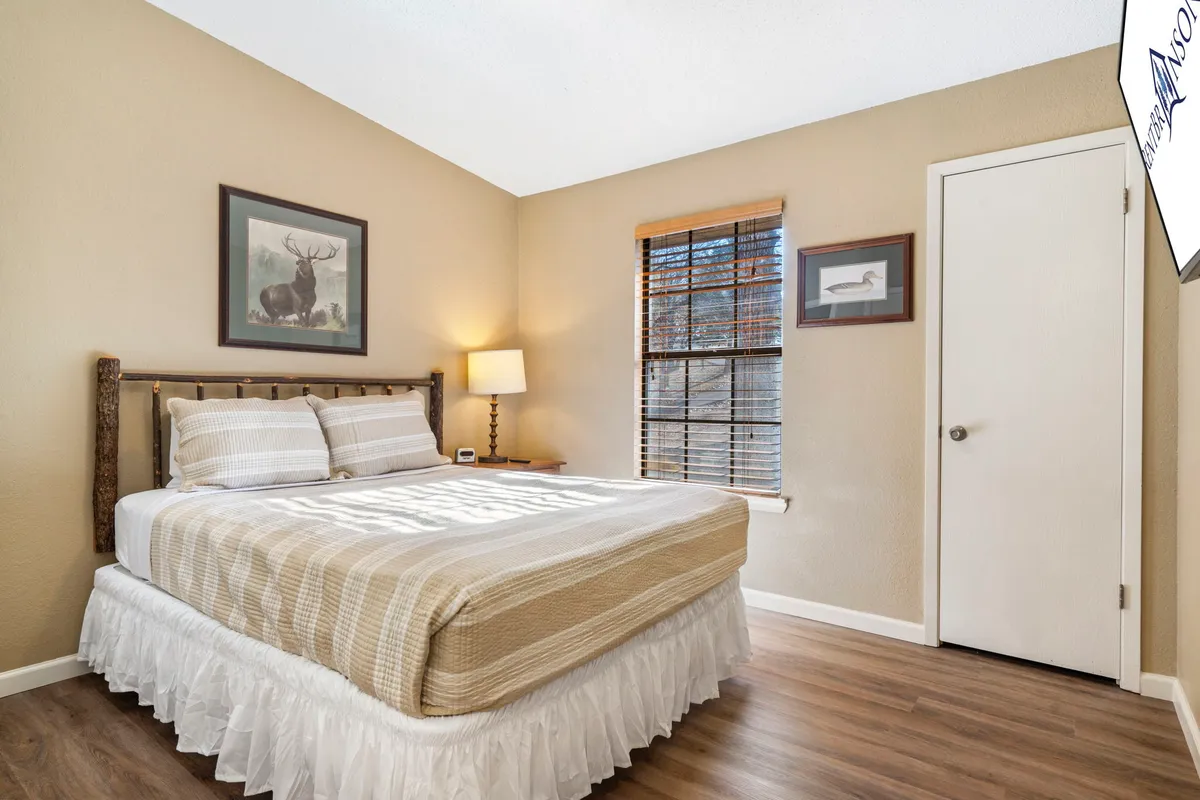 A serene bedroom with a neatly made bed, soft lamp lighting, a large window letting in natural light, and tasteful artwork on the wall. The space is uncluttered and exudes a calm, relaxing atmosphere, ideal for unwinding after a day of activities.
