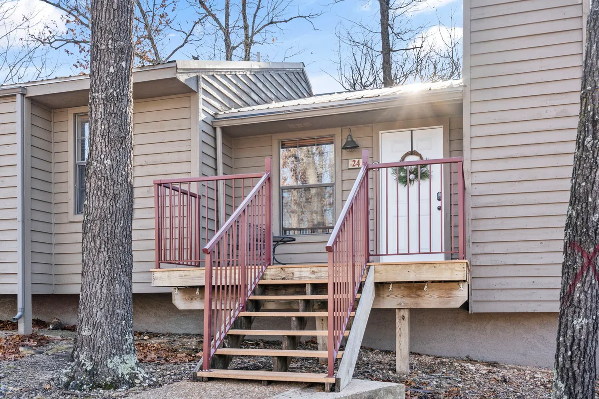Wooden steps leading up to a porch with a red railing and a welcoming front door adorned with a wreath. The porch is decorated, hinting at seasonal celebrations and a lived-in, inviting atmosphere. Trees can be glimpsed in the background, connecting the porch to its natural surroundings.